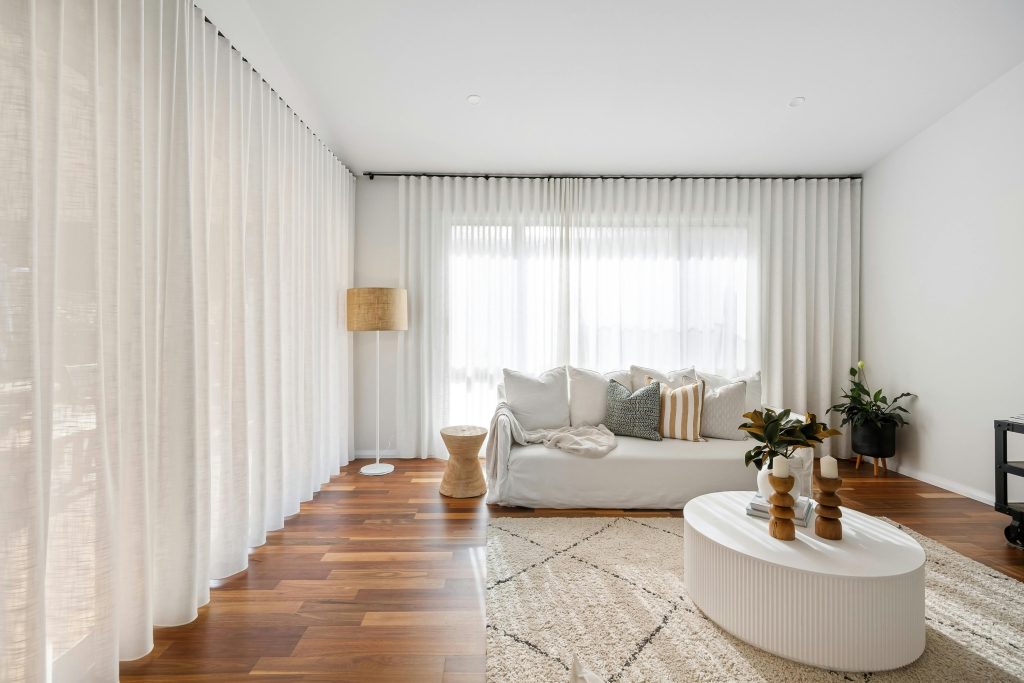 A bright living room with sheer window treatments, a white sofa, assorted pillows, a round coffee table, and a potted plant on the carpeted floor.