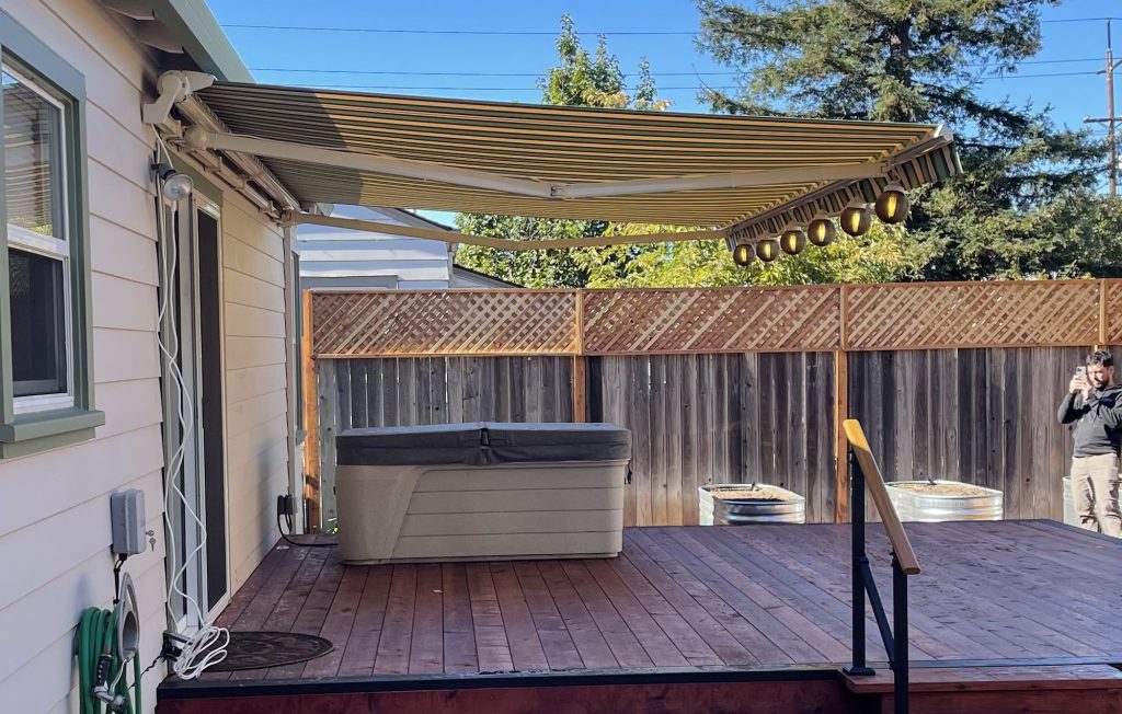 A wooden deck with a hot tub under a striped retractable awning, adjacent to a white house. A person stands by the fence, taking a photo. Trees and clear sky in the background.